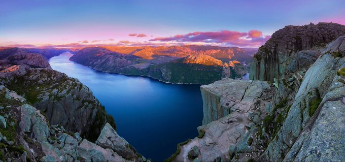 A scenic panoramic landscape photograph of a deep blue lake or fjord nestled between rocky mountains under a pink and purple sunset sky.