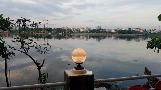 A scenic view of a calm river or lake reflects a cloudy sky and a distant shoreline dotted with buildings and a construction crane, with a spherical white lamp mounted on a post in the foreground.