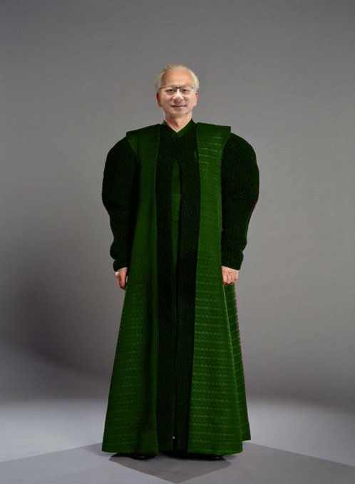 Man with glasses wearing an oversized green judicial-style robe, standing against a gray studio background.