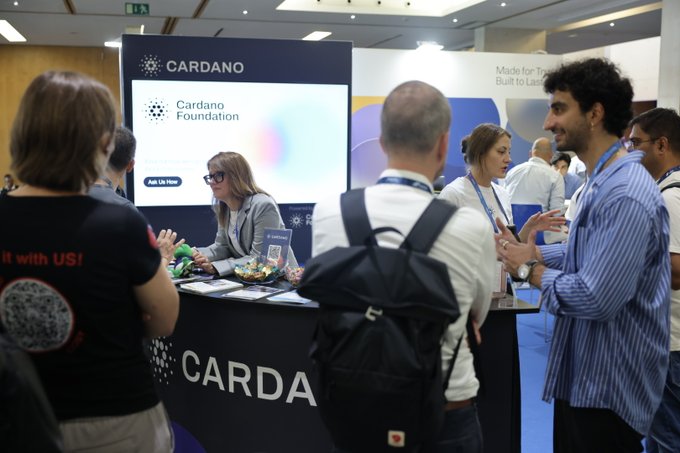 Attendees gather at the Cardano Foundation booth at what appears to be a blockchain conference, with staff engaging visitors in conversation around a branded display featuring the "CARDANO" name and logo.