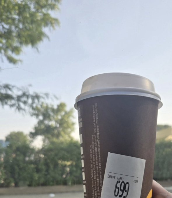 A hand holds a dark brown McDonald's drink cup with a white lid against a backdrop of trees and sky. The label reads 'DRIVE-THRU 699' and mentions McDonald's trademarks from 2013-2024.