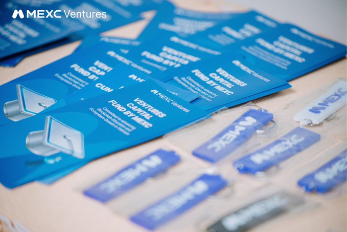 Blue promotional materials and keychains for 'MEXC Ventures' are laid out on a wooden surface, advertising their Fund B1.