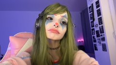 A person with green hair wears a painted mask over their eyes and nose, along with headphones, in a room with purple lighting and a photo-covered wall.