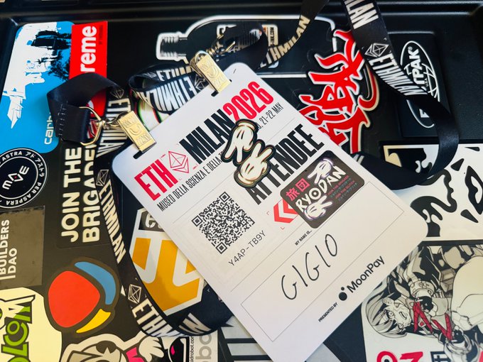 urn of water, white paper with black text and logos, red and black keychains, and a black case with stickers are scattered on the table.