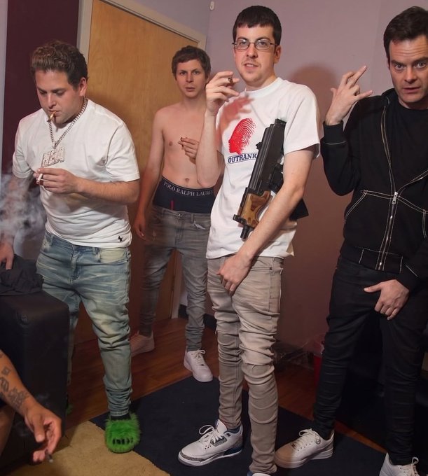 Four young men pose together indoors; one wears a white 'OUTBANK' shirt and holds a firearm, another smokes, and one sports bright green furry Crocs.
