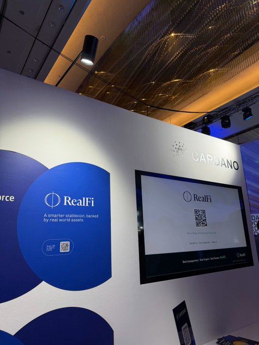 An exhibition booth for 'RealFi' by Capuano, with text describing it as 'A smarter stablecoin, backed by real world assets,' featuring a QR code on a digital screen.
