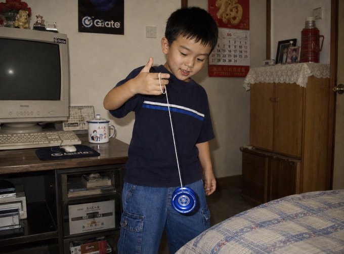 The image features a young boy holding a blue toy wind-up toy, which appears to be a yo-yo or a similar object that can be wound up and played with. The boy seems to be enjoying himself as he plays with his toy in front of a computer desk. There is also a TV present in the scene, suggesting that this might be a living room setting where people gather for various activities.