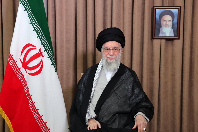 An elderly cleric with a white beard, wearing a black turban and clerical robes, sits in front of an Iranian flag, with a framed portrait of another bearded cleric hanging on the wall behind him. The image connects to the post's claim about cheap Bitcoin mining in Iran by visually representing the country's religious and political leadership, under whose governance the low energy costs enabling profitable mining exist.