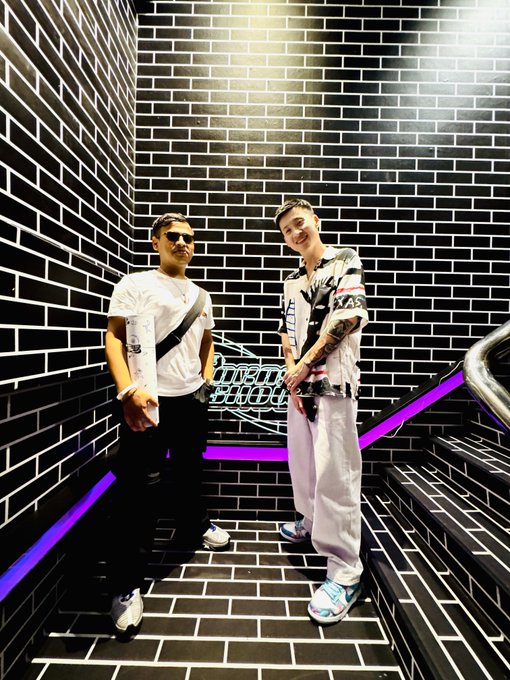 Two stylish friends pose on a neon-lit staircase against black brick walls, with one wearing a white "XANS" shirt featuring Japanese characters. The vibrant setting and their close friendship reflect the post's heartfelt birthday tribute celebrating their bond within the ₿-R4ted Community.