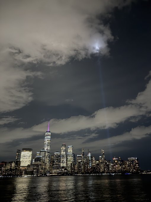 A nighttime view of the New York City skyline, including One World Trade Center, with a beam of light shining up through dramatic clouds over the water.