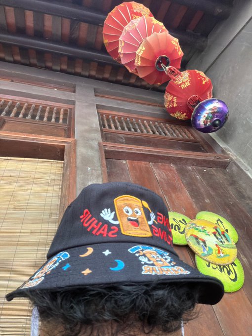 A person poses wearing a "ZAP ZAP" graphic bucket hat and yellow flip-flops, with "tung tung tung" resonating alongside the loud red Chinese lanterns hanging overhead in a traditional wooden building.