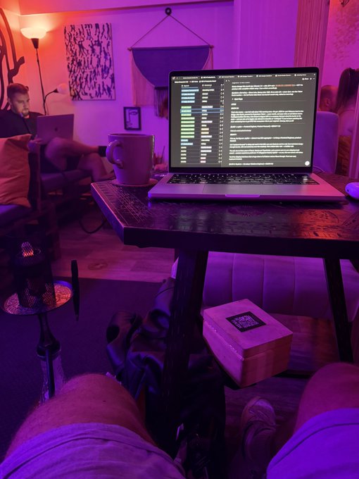 A person works on their laptop displaying code in a dimly lit room with purple lighting, accompanied by a coffee mug and a wooden box, evoking a late-night work or coding session that aligns with the post's mention of a packed week featuring a Solflare livestream, Solana Summit, and a live podcast taping.