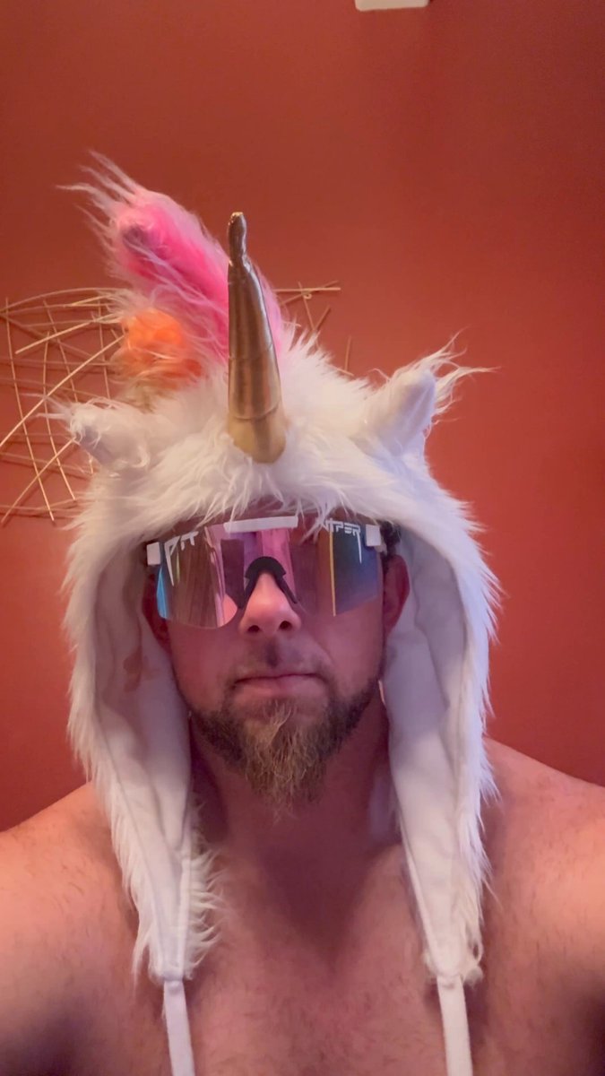 The image features a man wearing a white unicorn hat with pink and orange accents, sunglasses, and no shirt. The man appears to be posing for a photo or selfie, possibly sharing his unique style choice with others.
