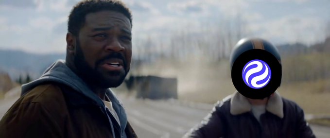 A man with a beard wearing a brown jacket looking surprised, standing next to a person in a motorcycle helmet with a purple logo obscuring their face.
