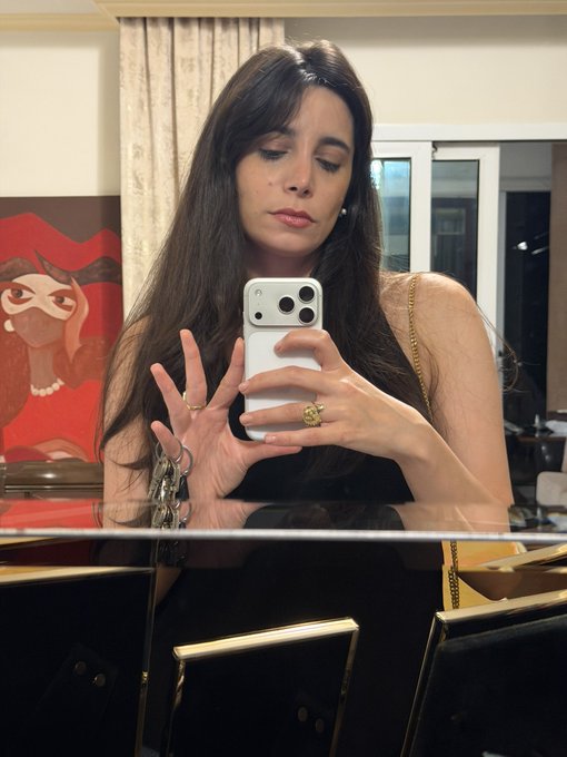 A mirror selfie of a woman with long dark hair holding a phone, wearing a black outfit with gold chain straps and rings.