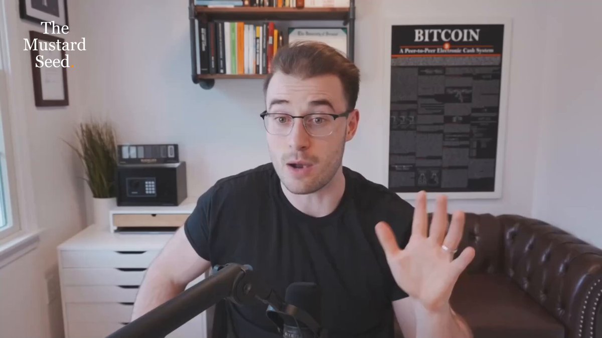 The image shows a man wearing glasses sitting at a desk with a microphone in front of him. He appears to be giving a speech or presentation, possibly related to Bitcoin or the stock market. The background features a bookshelf filled with various books, suggesting that he might be discussing financial topics or sharing insights about investing and cryptocurrency markets.