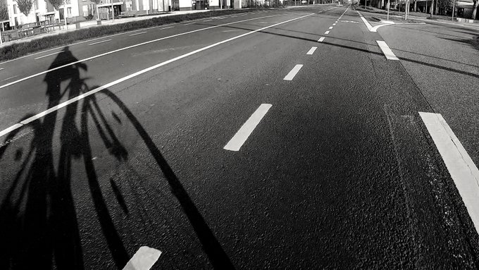 A black and white photograph showing the elongated shadow of a cyclist cast across an empty, multi-lane paved road.