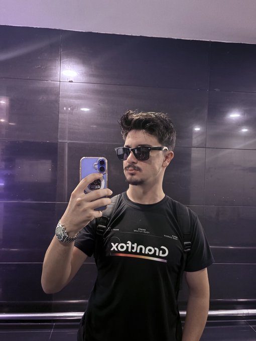 A young man with sunglasses takes a mirror selfie in an elevator, wearing a black "Caratlax" t-shirt and a silver watch, with a backpack strap visible over his shoulder. The caption suggests he's referencing his partner, pairing the stylish, mirror-selfie vibe with a personal "wake up" message.
