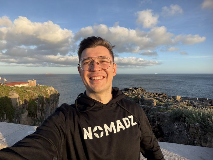 A smiling young man wearing glasses and a black 'NOMADZ' hoodie takes a selfie with a scenic coastal background featuring cliffs, ocean, and a cloudy sky.