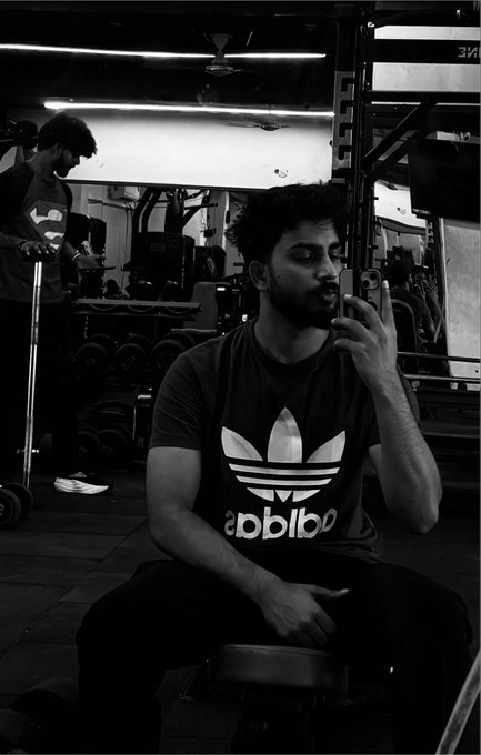 A black and white photograph captures a muscular man wearing an Adidas t-shirt sitting in a gym, gesturing with his hand while speaking, with another person using a barbell in the background among gym equipment.