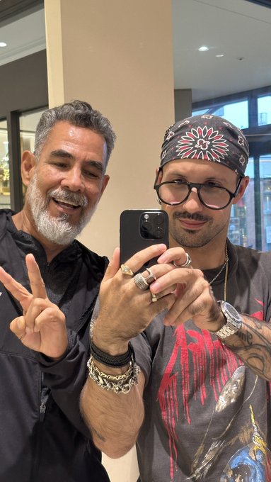 Two men take a mirror selfie with a smartphone; the older man with a gray beard makes a peace sign, while the younger man wears glasses and a bandana.