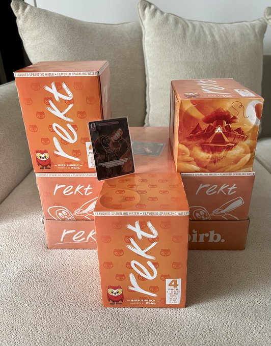 Multiple orange boxes of "rekt" flavored sparkling water in "BIRB BUBBLY" peach and maracuja flavor (4 pack) are stacked on a beige couch, with a delivery card on top, illustrating the timely arrival of @rektdrinks ($REKT) products as promised.
