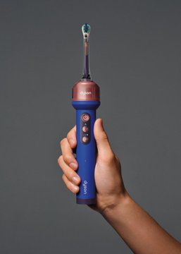 A hand holds a blue and rose gold electric toothbrush against a gray background, displaying a toothbrush head at the top and control buttons on the handle.