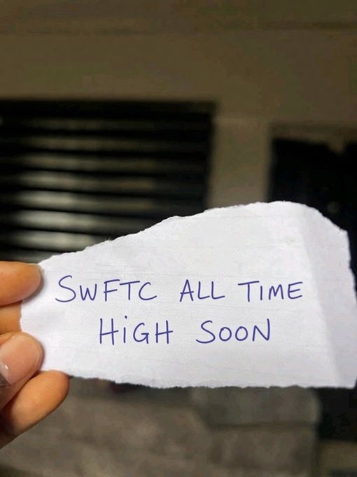 A hand holding up a torn piece of paper with handwritten blue text that reads 'SWFTC ALL TIME HIGH SOON,' referring to the SWFTC crypto ticker.
