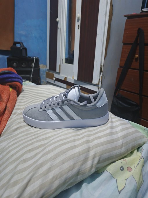 A gray and white Adidas sneaker with the iconic three-stripe design rests on a striped pillow in a bedroom, matching the caption's description of original Adidas shoes purchased affordably on eBay and shipped to Venezuela.