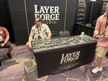 A vendor sits at a 'LAYER FORGE STUDIO' booth, likely at a gaming convention, with a table covered in merchandise and a customer browsing on the right.