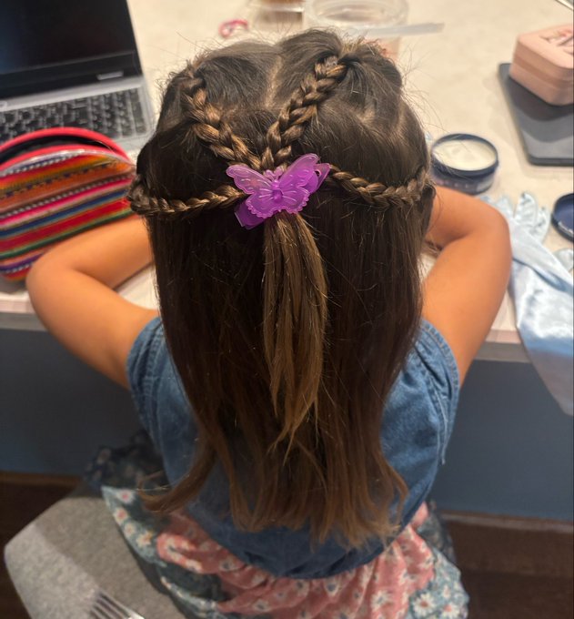A young girl seen from behind sitting at a desk, wearing a denim top with intricately braided hair adorned with a purple butterfly hair clip.