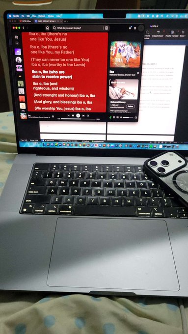 A MacBook Pro sits on a bed with a music streaming app open displaying lyrics including 'Iba o, Iba (there's no one like You, Jesus)', 'Iba o, Iba (who are able to receive power)', and 'Iba o, Iba (the most righteous, and wisdom)', with an iPhone resting on the keyboard.
