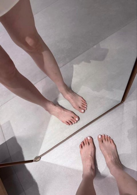 A pair of bare feet stands on a tiled floor in front of a mirror, with their reflection clearly visible in the glass. The image captures a quiet, introspective moment that aligns with the theme of "reflection" through its literal mirror imagery.