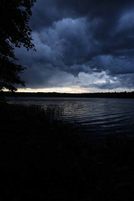A dark, moody photograph of a lake at dusk under heavy storm clouds, with the silhouette of trees framing the left side.