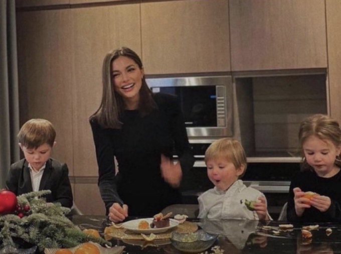 A woman in a black outfit laughs while standing in a modern kitchen with three children seated around a counter that has plates of food and a Christmas decoration with a red ornament.