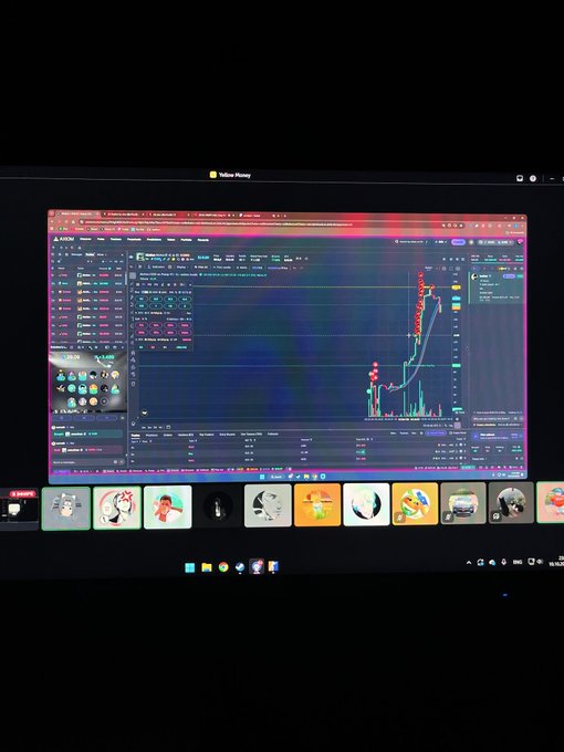 A monitor displays a TradingView cryptocurrency chart showing a sharp upward price spike with candlestick patterns and volume indicators, with a taskbar of social media icons at the bottom.