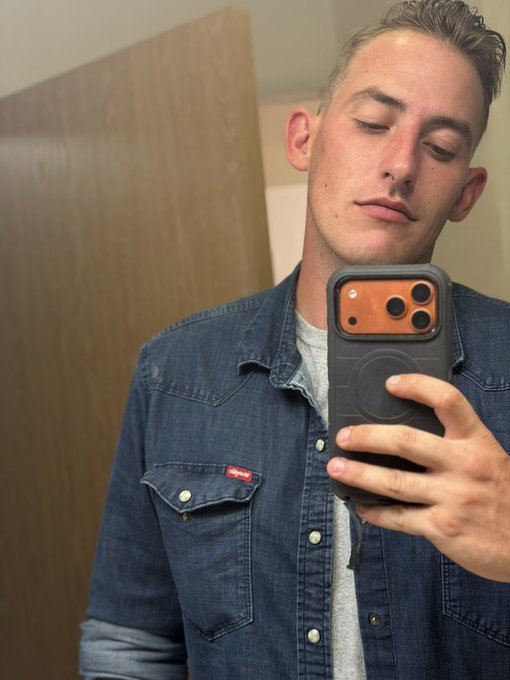A man taking a mirror selfie in a denim Wrangler shirt (with the visible red "Wrangler" logo tag) while holding his iPhone, eyes closed with a calm expression — projecting the relaxed, confident "GM" Friday energy the post channels.