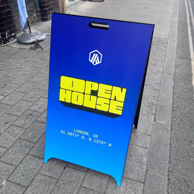 A blue A-frame sidewalk sign on a London street advertising an 'OPEN HOUSE' event with coordinates 51.5072° N, 0.1276° W.