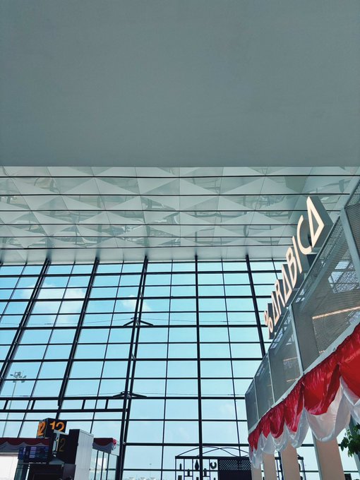 The interior of a modern airport terminal features floor-to-ceiling glass windows, with red and white draped decorations and an "Emirates" sign visible on the right, suggesting a festive setup for Coinfest. A display showing "2.12" is seen on the lower left, possibly indicating the event date or time.