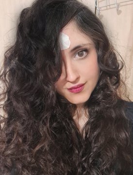 A selfie portrait of a young woman with long curly dark hair and red lipstick, wearing a small white heart-shaped bandage on her forehead, posing in what appears to be a bathroom setting.
