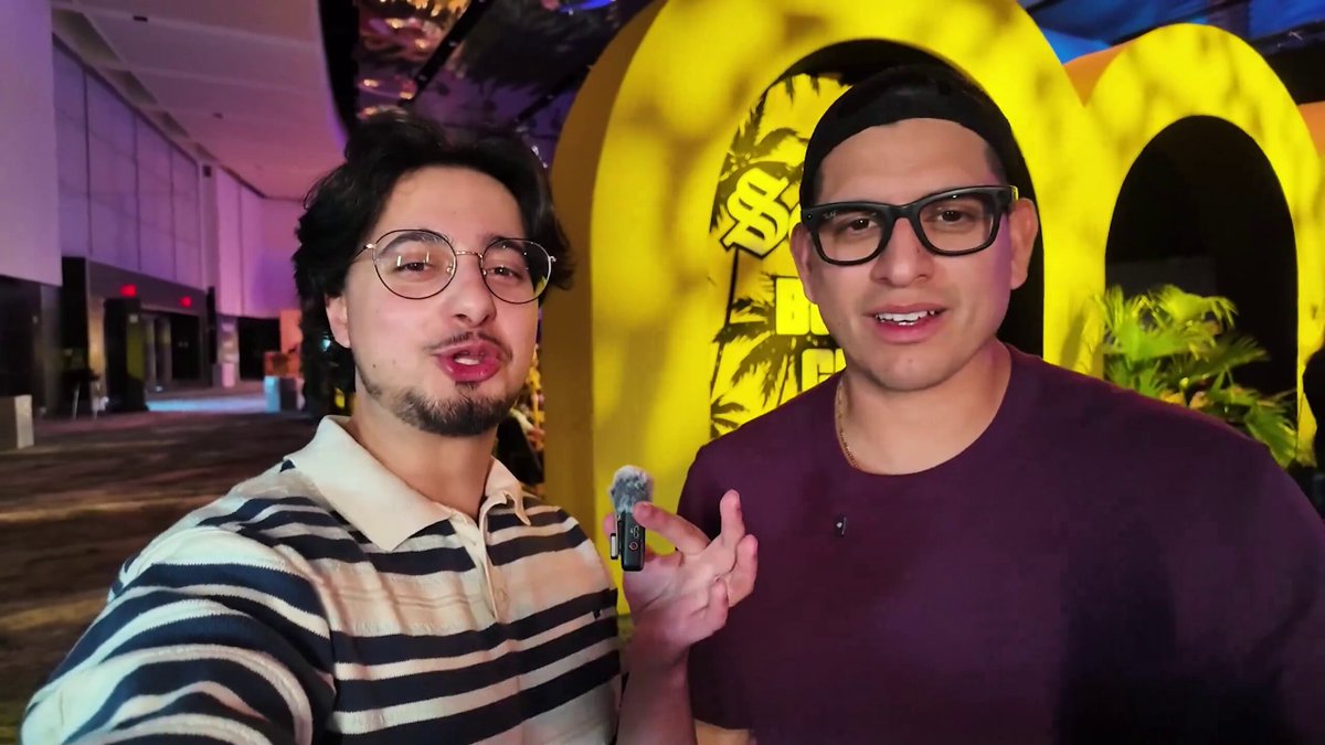 The image features two men posing for a picture together with one of them holding up an item that appears to be a Pokemon card. The man holding the Pokemon card has a large smile on his face while the other man looks at him, possibly admiring or discussing the card. The background shows a hallway and a door, suggesting they are in a public space like a mall or a gaming convention.