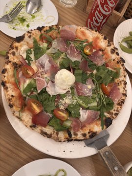 A pizza topped with prosciutto, arugula, tomatoes, and a ball of mozzarella cheese served on a white plate, with a Coca-Cola bottle visible in the background.