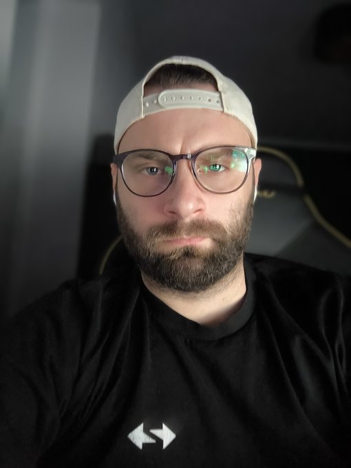 A selfie of a bearded man wearing glasses and a backward white cap, with a deadpan expression that matches the post's exhausted mood about waiting for $ZK to hit $2 on day 703, his black shirt displaying small white arrows.