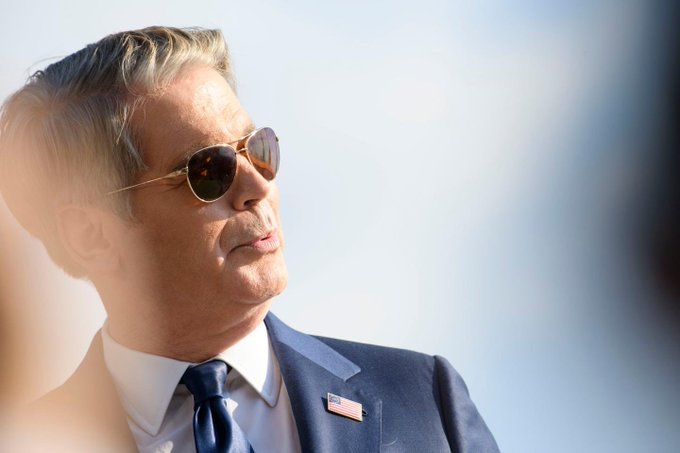 A close-up portrait of an older man with grey hair wearing aviator sunglasses, a dark blue suit, and an American flag pin on his lapel, looking off into the distance against a clear blue sky.