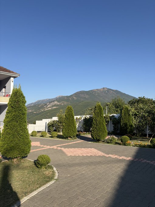 A paved courtyard with manicured shrubs and trees overlooks a vast mountain range under a clear blue sky.