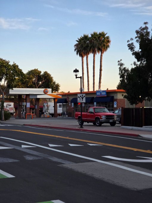 A 76 gas station at dusk with a red pickup truck parked in front, palm trees in the background, and an empty road in the foreground.