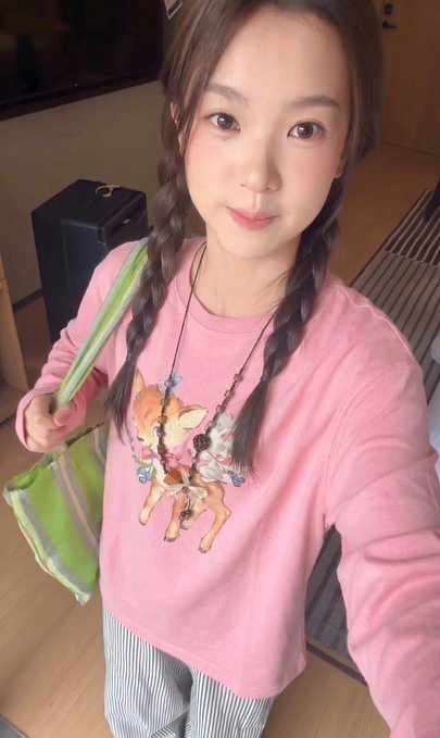 A selfie of a young woman with twin braids wearing a pink sweatshirt with a deer graphic, holding a green tote bag.