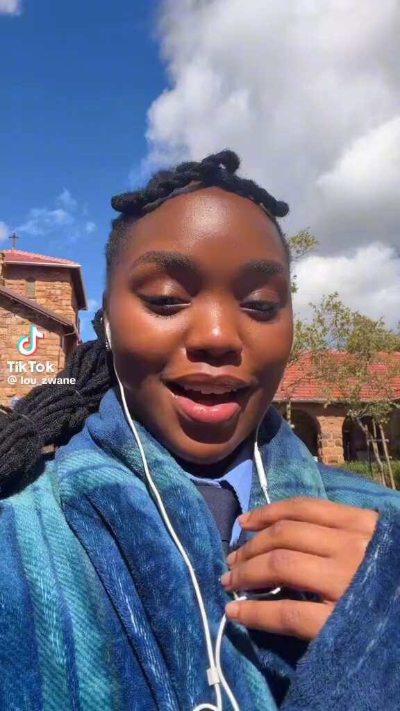 The image features a woman with dreadlocks wearing a blue plaid blanket, smiling at the camera while holding a white earbud. The background includes a house and a sign that reads "TikTok 2.5".