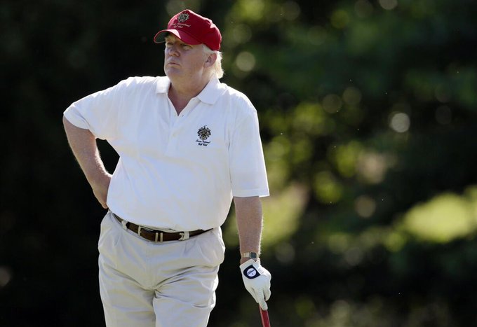 Donald Trump stands on a golf course wearing a red 'MAGA' cap and a white polo shirt, holding a golf club while wearing a white glove.