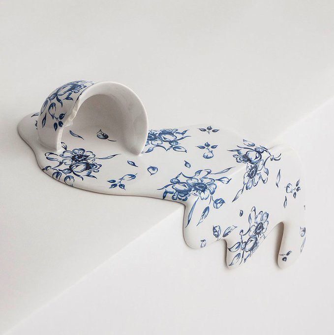 A white ceramic sculpture with blue floral patterns depicting a melting cup pouring into a puddle that forms an animal shape, resembling traditional blue-and-white porcelain.